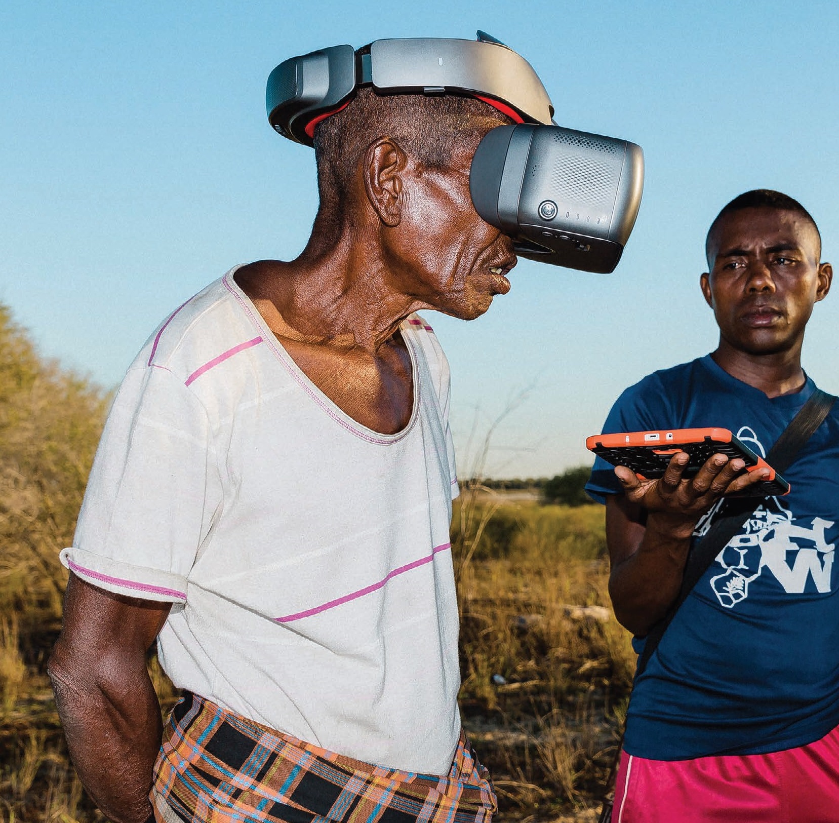 Blue sky, a man has a virtual reality mask, another man holds a tablet while looking at the first man..